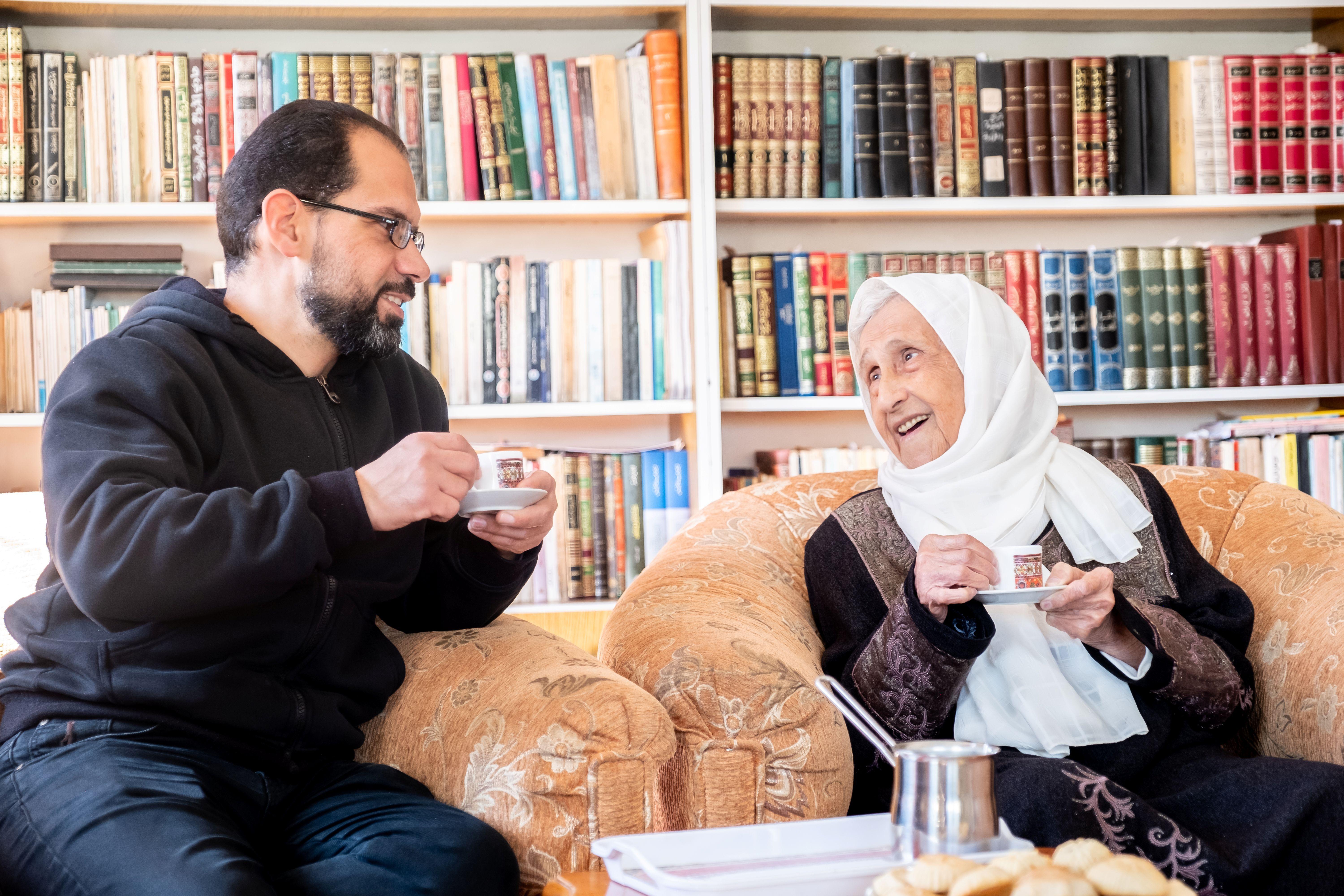 رجل وامرأة مسنة ترتدي حجاباً أبيض يجلسان في غرفة معيشة أمام مكتبة، ويتبادلان الحديث وهما يحملان فنجاني قهوة
