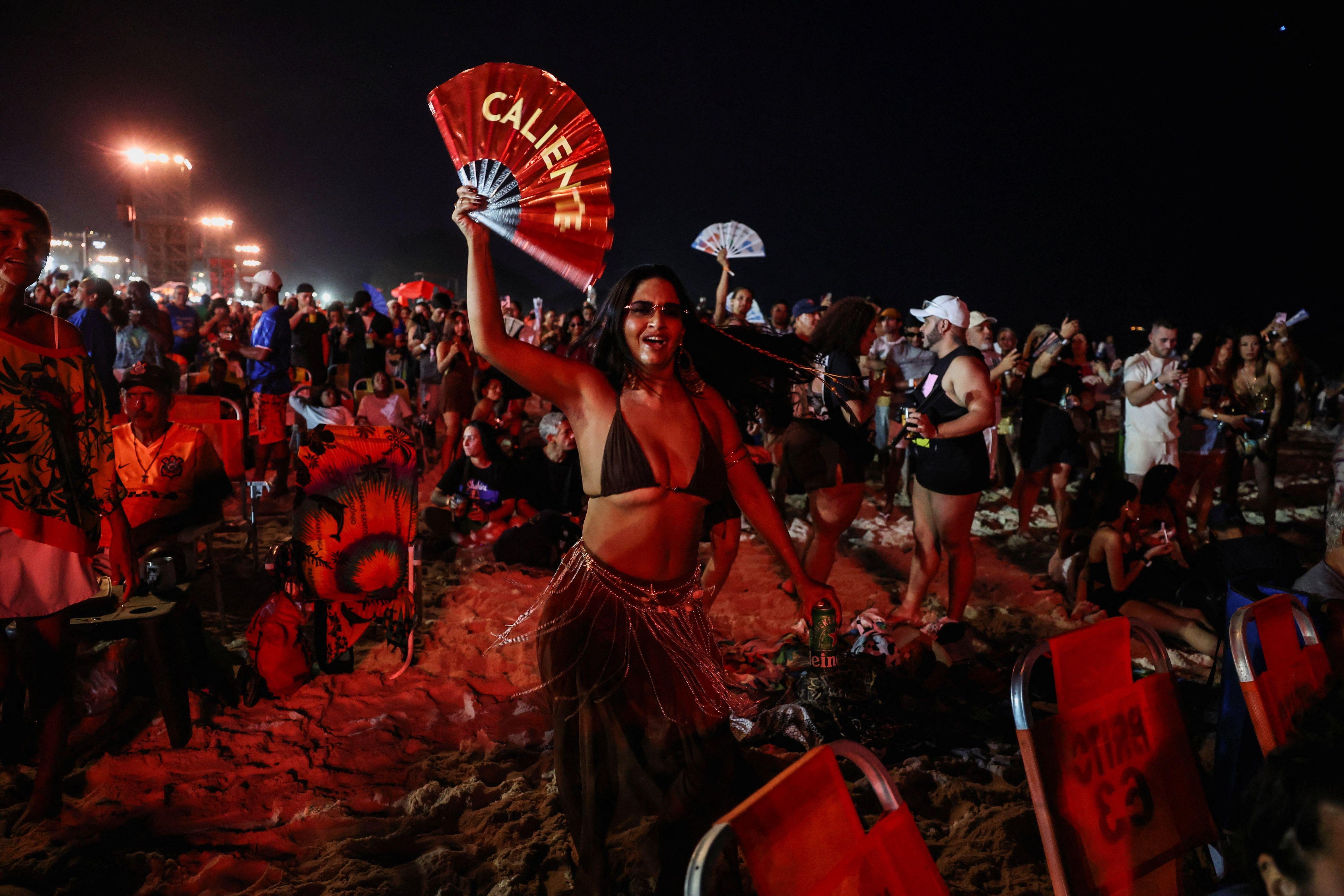 Mulher segura leque com a palavra caliente. Ela est&aacute; na praia, &agrave; noite. Muita gente atr&aacute;s. 
