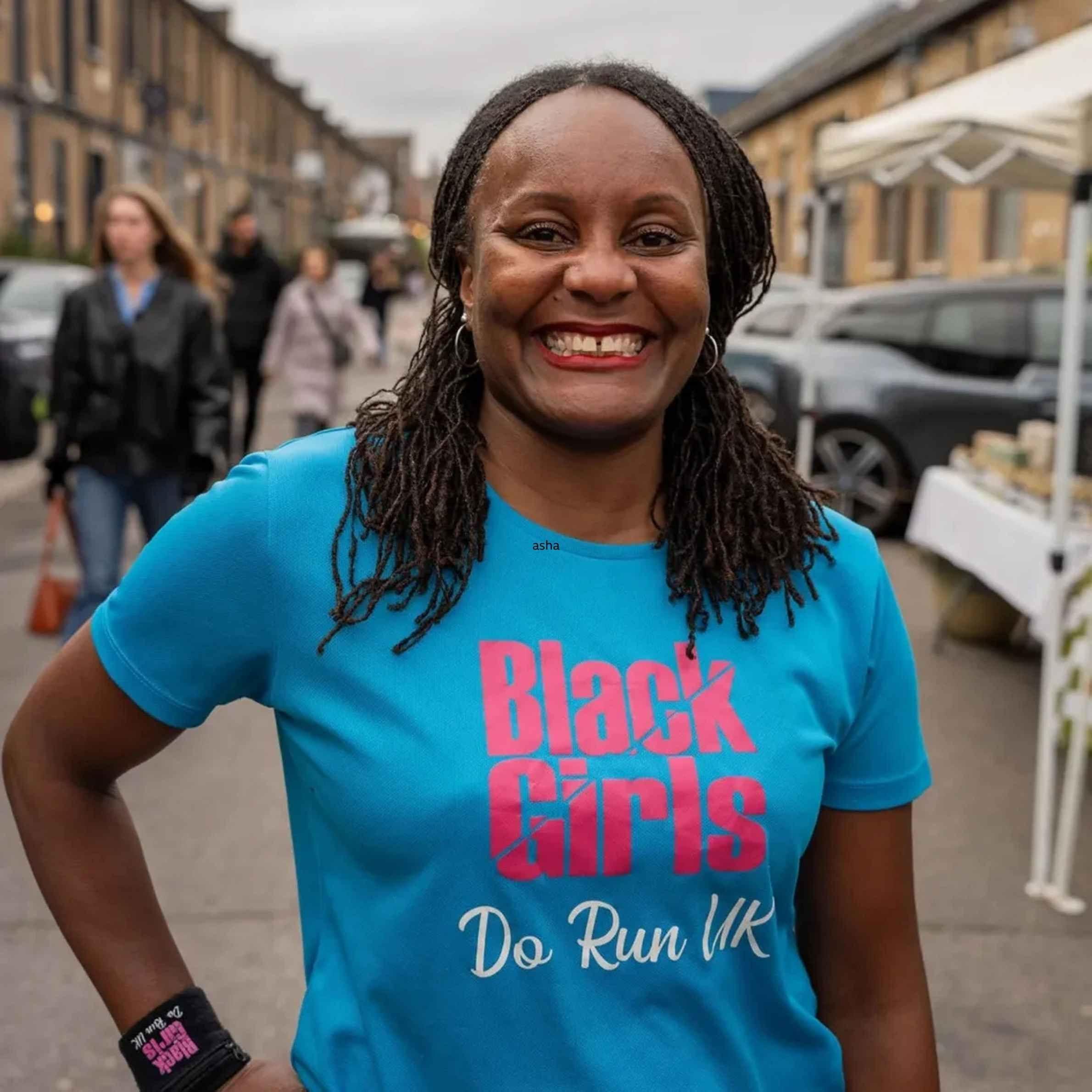 Tasha beams at the camera with a hand on her hip. She stands outside on the street, and in the blurry background you can make out some passersby and a stall. Her hair is braided, and she wears a blue t-shirt, with pink writing that reads 'Black Girls Do Run UK.'