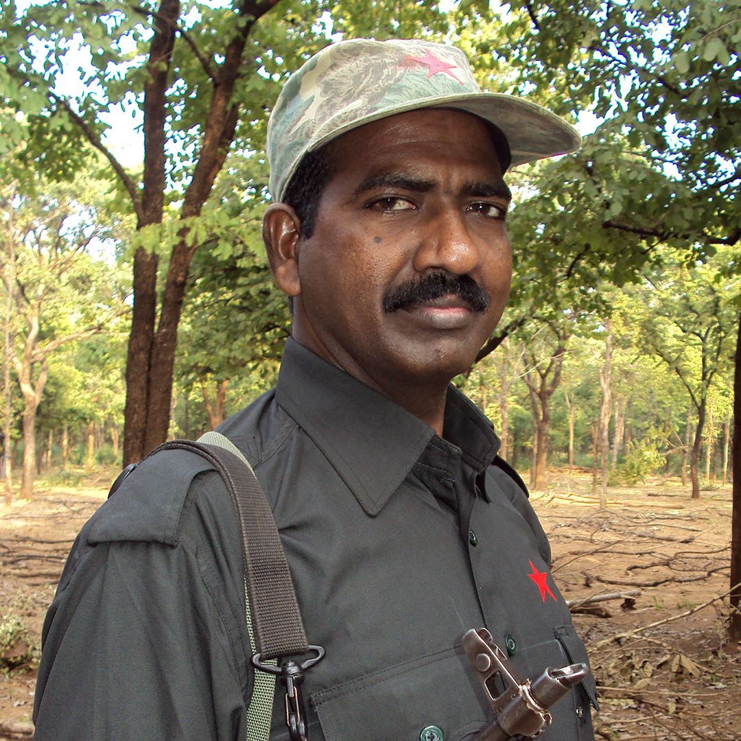 Seorang pria mengenakan kemeja abu-abu kehijauan gelap dengan bintang merah di dadanya. Ia berkumis dan mengenakan topi bisbol kamuflase bergambar bintang merah pudar di bagian depannya. Ia menenteng pistol di bahunya. Ada pepohonan di latar belakang.