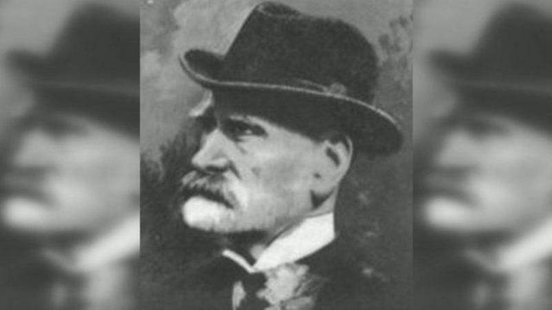 Foto en blanco y negro de Cobb Morley, de perfil, con un sombrero, camisa de cuello alto, corbata antigua y chaqueta, estilo victoriano. lleva barba y bigote. 
