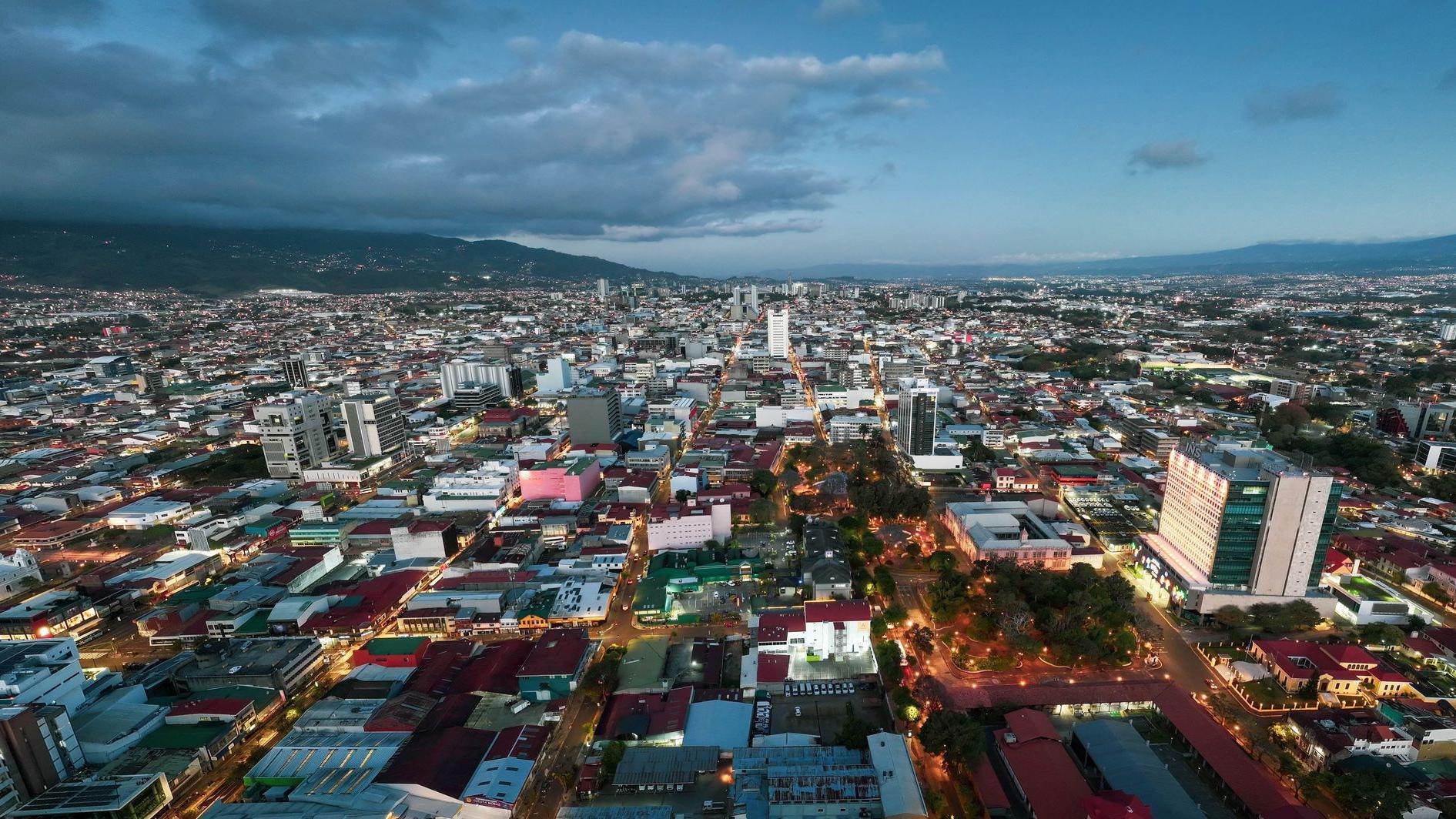 Una foto aérea de San José, Costa Rica. Parace un final del atardecer, se ven ya algunas luces encendidas en parques y avenidas, con una montaña de fondo.
