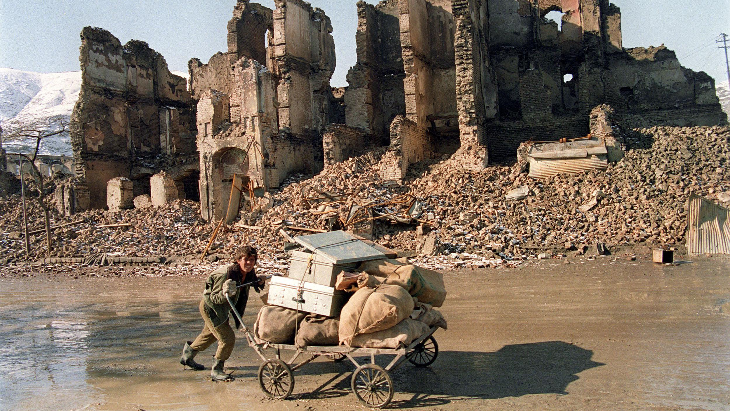 Un joven afgano empuja una carreta cargada de sacos y cajas por una calle embarrada. Detrás de él se ve un edificio destruido y escombros.
