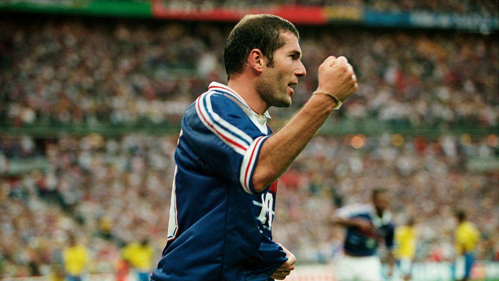 A imagem mostra um jogador da sele&ccedil;&atilde;o francesa comemorando de forma vibrante durante uma partida, com o punho cerrado e express&atilde;o de entusiasmo. Ao fundo, um est&aacute;dio lotado aparece desfocado, enquanto jogadores brasileiros podem ser vistos &agrave; dist&acirc;ncia, refor&ccedil;ando o contexto de um confronto internacional.