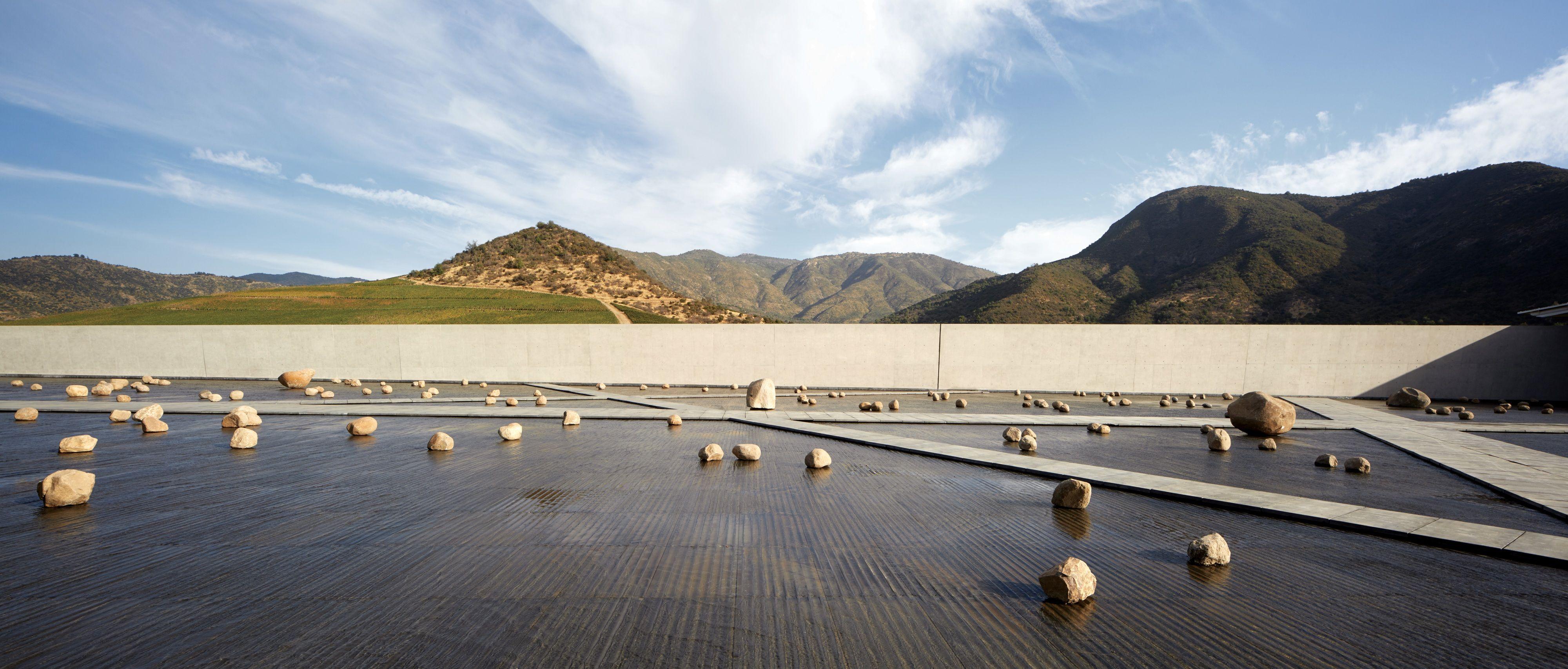 Patio con lámina de agua y rocas dispersas, enmarcado por un muro recto y un paisaje de montañas bajo un cielo despejado.