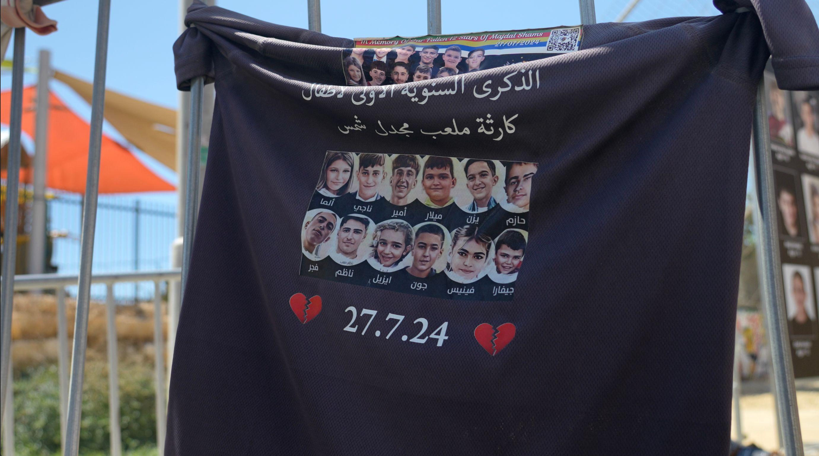 قطعة من قماش أسود مطبوع عليها صور وأسماء 12 طفلاً وقد علقت على سور معدني. وقد طبع أسفل قطعة القماش صورتان باللون الأحمر لقلبين مكسورين وتاريخ 27.7.24 باللون الأبيض.