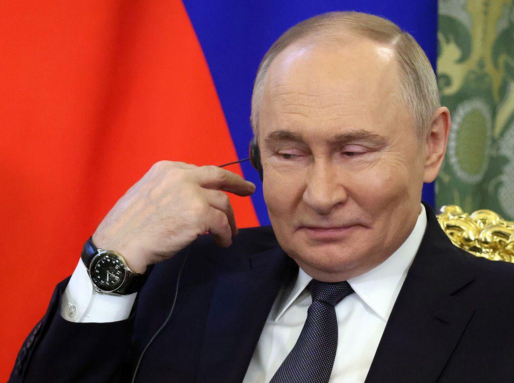 Vladimir Putin, con gesto sonriente, se lleva la mano derecha a la oreja derecha, en la que tiene un audifono. 