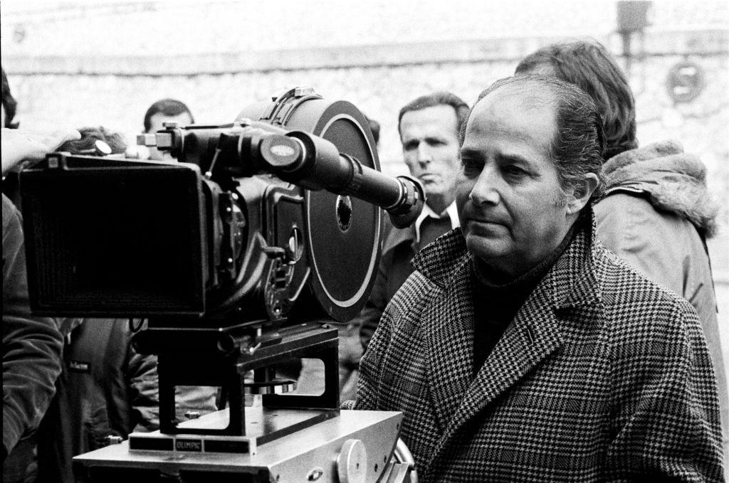 En una foto en blanco y negro, Gillo Pontecorvo está vestido con un abrigo y parado detrás de una cámara de cine de 35mm. Lo rodean otros miembros del equipo de filmación