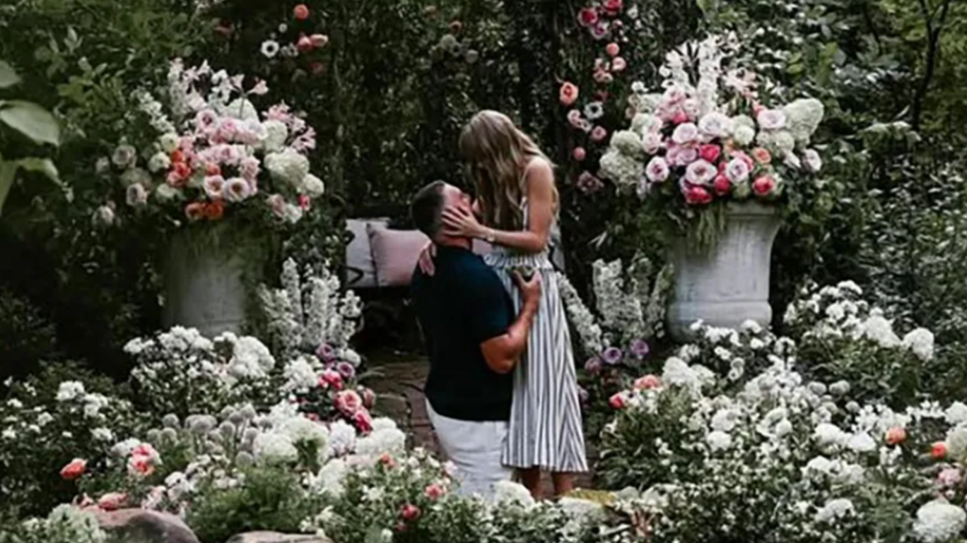 En un jardín frondoso con flores blancas y rosas, un hombre vestido de pantalón blanco y una camisa oscura de manga corta se arrodilla frenta a una mujer rubia, vestida en un atuendo blanco de rayas negas y sin mangas que le toma de la cara con sus manos