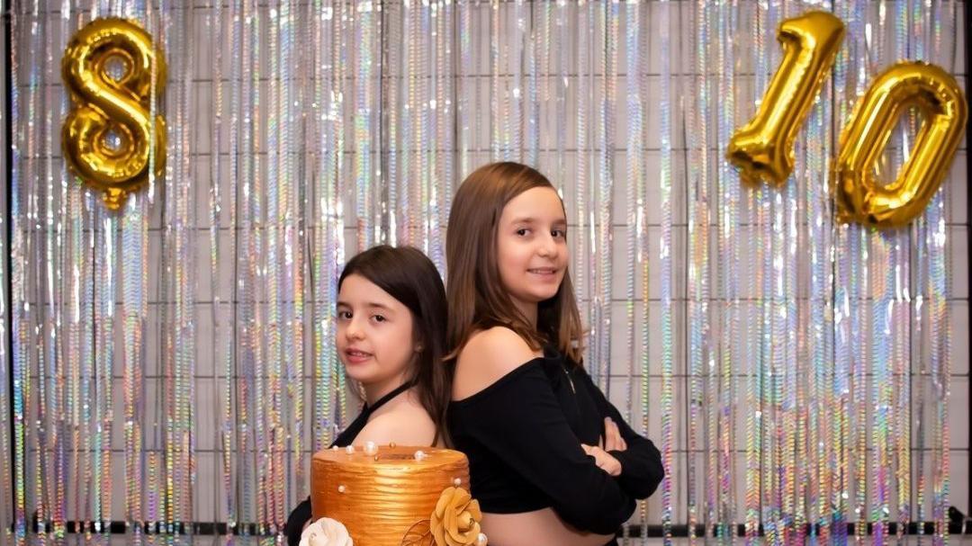 Beatriz and Lara Bernardi stand back to back behind a gold birthday cake. In the background are silver party decorations and two gold balloons - one says 8 and the other says 10.