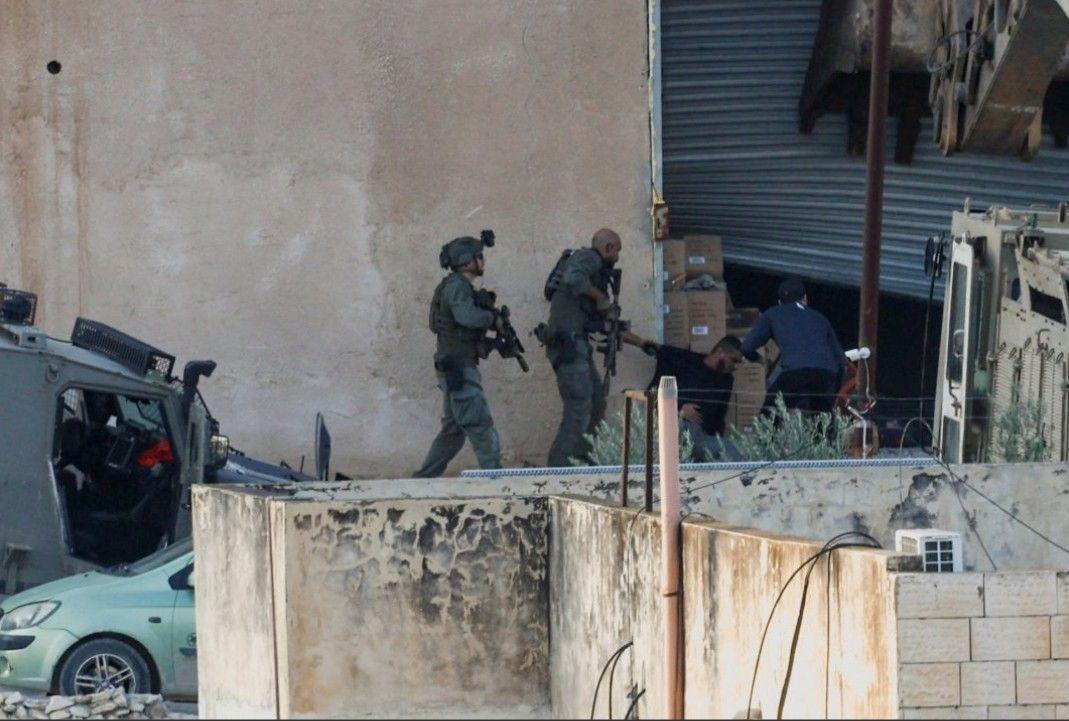 عنصران من القوات الإسرائيلية بزي عسكري يحملان السلاح، أثناء عملية جنين في الضفة الغربية المحتلة، يوجهان السلاح نحو شابين فلسطينيين يرتديان لباساً مدنياً. الصورة التقطت نهاراً.