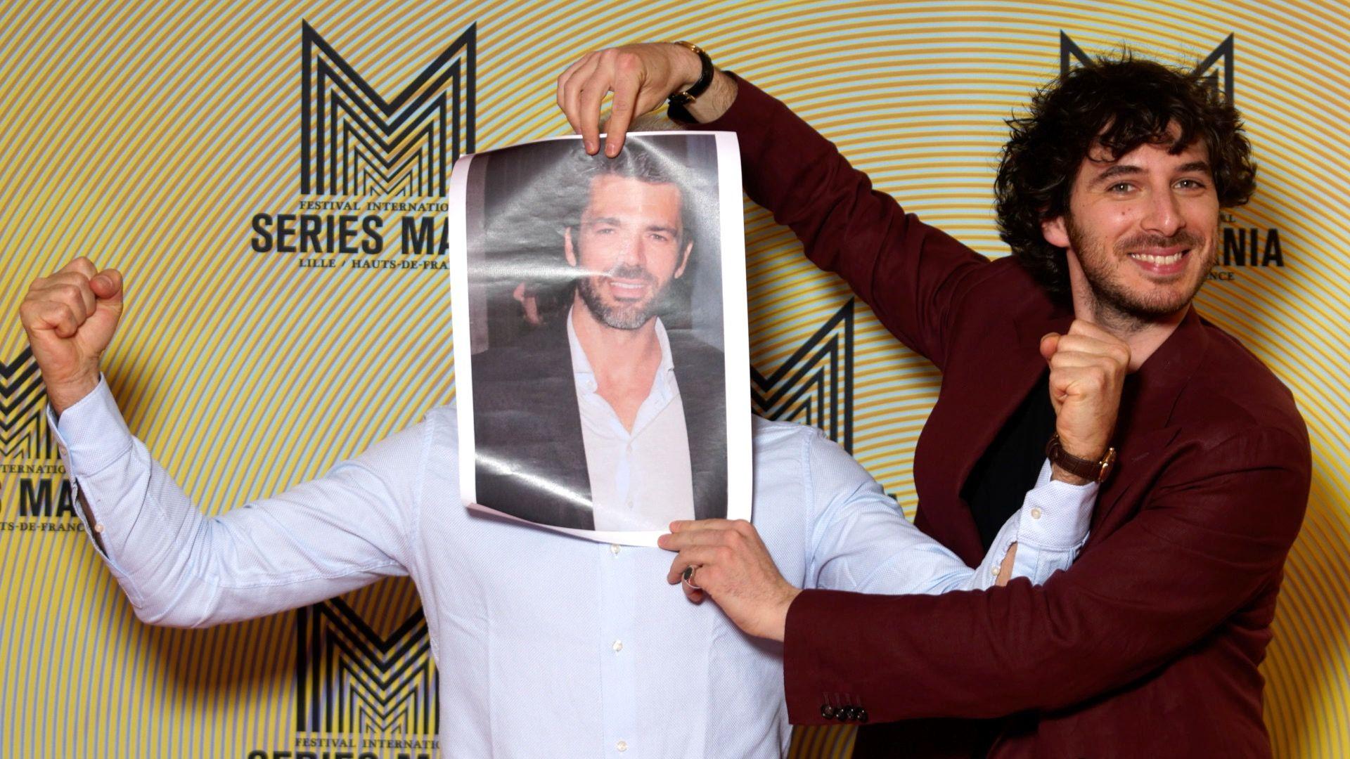 Actor Pierpaolo Spollon (right) holds a poster showing his RAI TV character over Pierdante Piccioni's face, as Piccioni flexes his arms and fists to indicate strength, at the Series Mania Festival, on 21 March 2022 in Lille, France