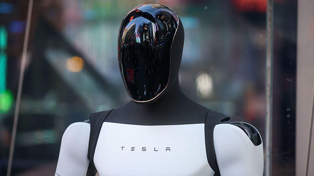 O robô Tesla Optimus. Ele tem forma humana — é possível ver sua cabeça e ombros. A cabeça é preta, brilhante e reflexiva, enquanto o torso e os braços são brancos. No centro do peito, está escrito “Tesla”. 