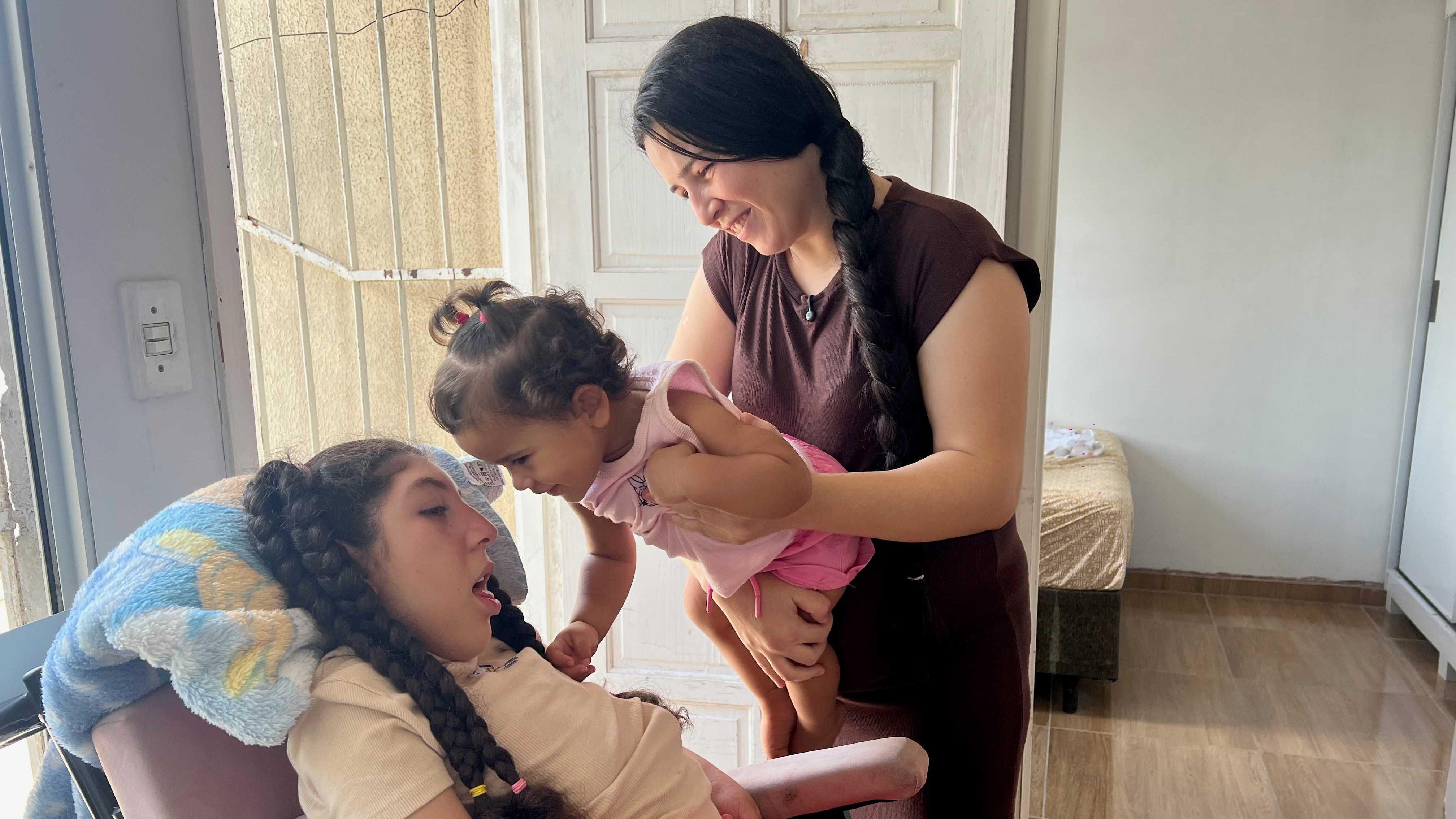 Inka segura Ester no colo enquanto ela toca o rosto da irm&atilde; Sophia, que est&aacute; em uma cadeira de rodas