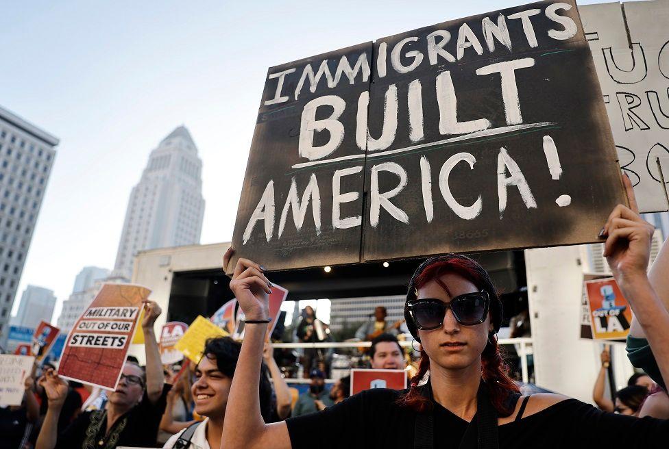 Una manifestante con lentes oscuros y una pancarta que dice "Immigrants built America!"
