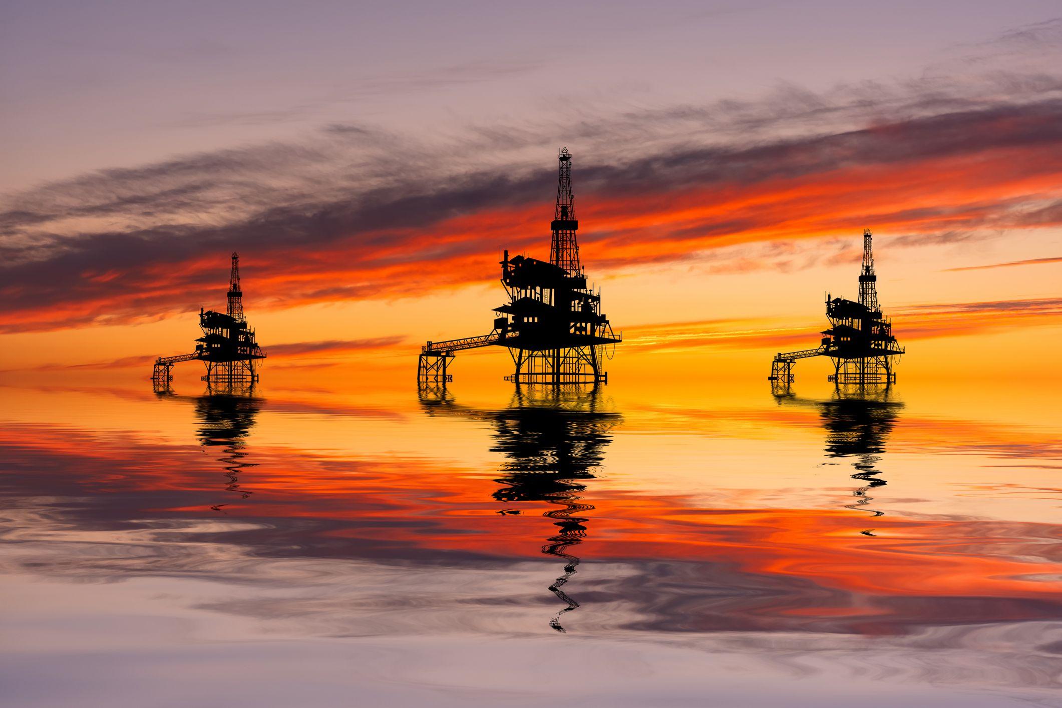 Tr&ecirc;s plataformas de petr&oacute;leo ao p&ocirc;r do sol. O c&eacute;u e o reflexo na &aacute;gua apresentam tons de laranja escuro e laranja claro, respectivamente. Os reflexos das plataformas na &aacute;gua formam ondas.