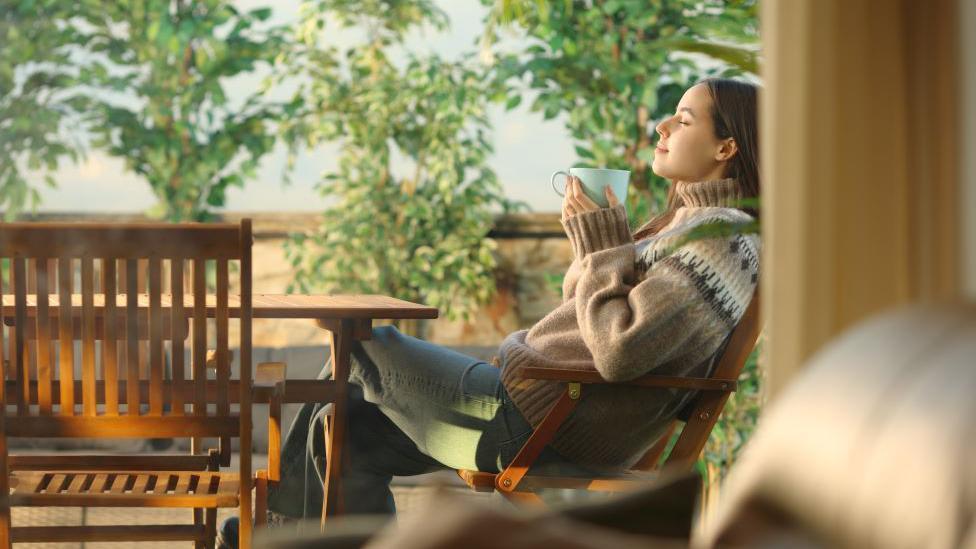 Žena u džemperu sedi na terasi ispred kuće, drži piće u šolji i opuštena je sa zadovoljnim izrazom lica.