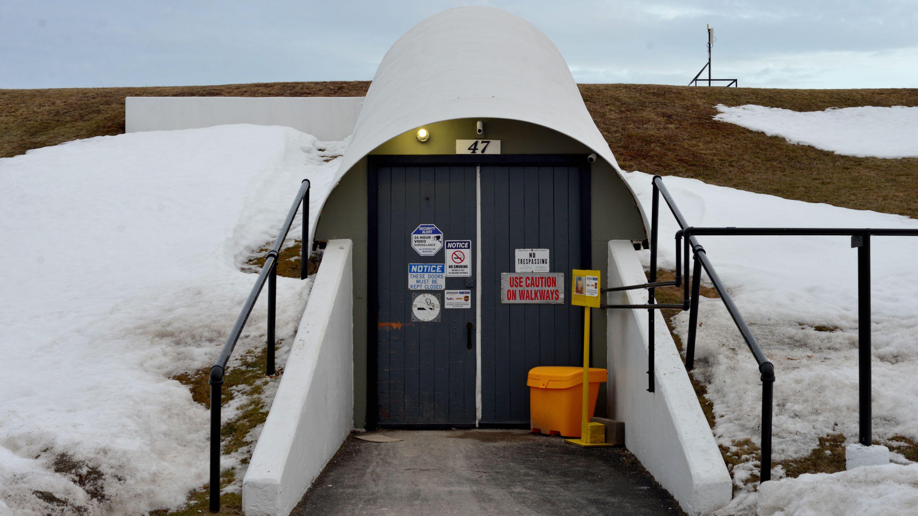 Dos puertas negras con señales de advertencia construidas en una colina cubierta de nieve, con algo de hierba seca asomando. Están cubiertas por un techo blanco en forma de arco y un camino, con paredes de hormigón blanco, está pavimentado frente a ellas.