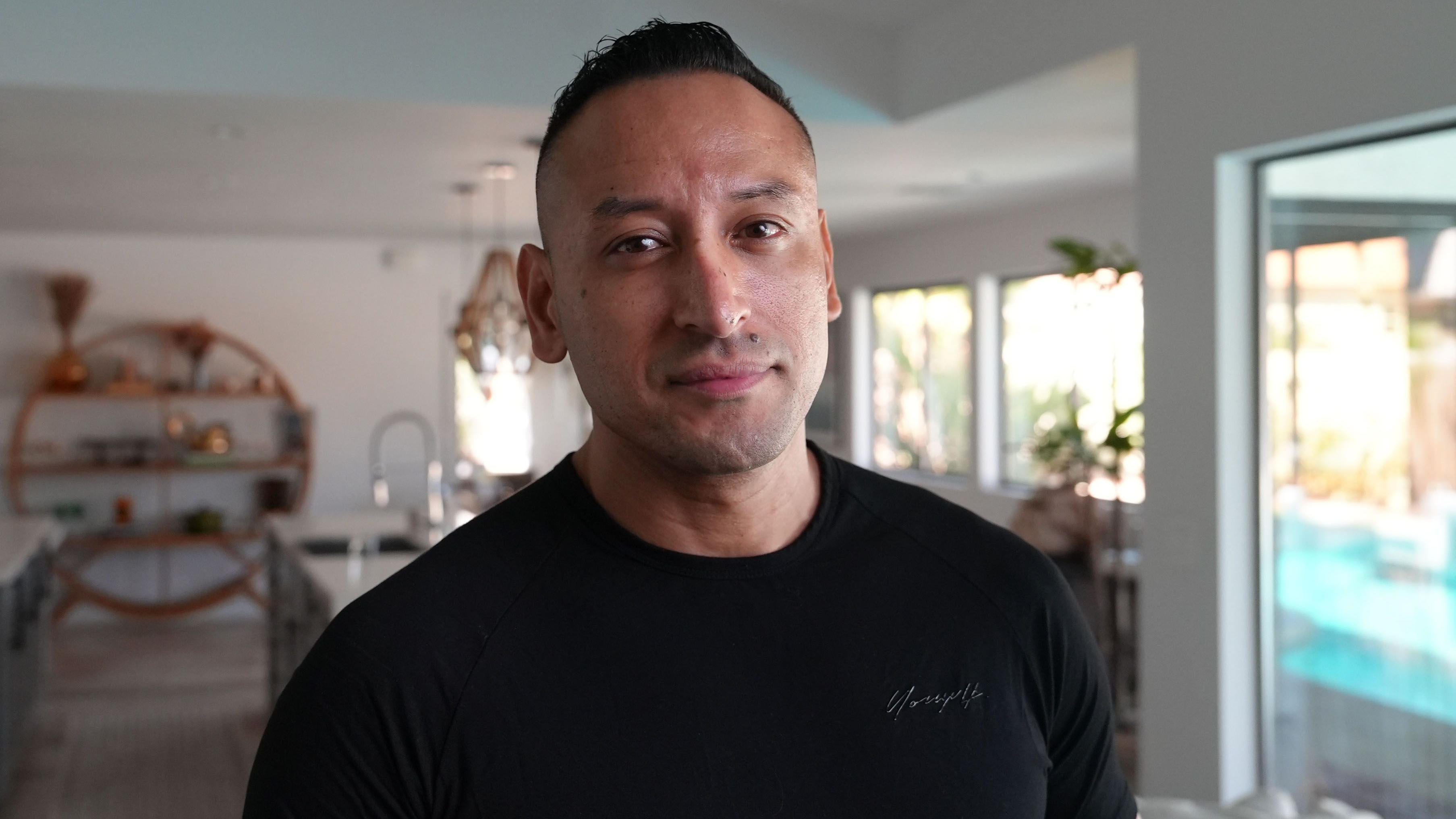 Tony Marquez, de pé, no que parece ser a sala de uma casa. Ele usa uma camiseta preta.