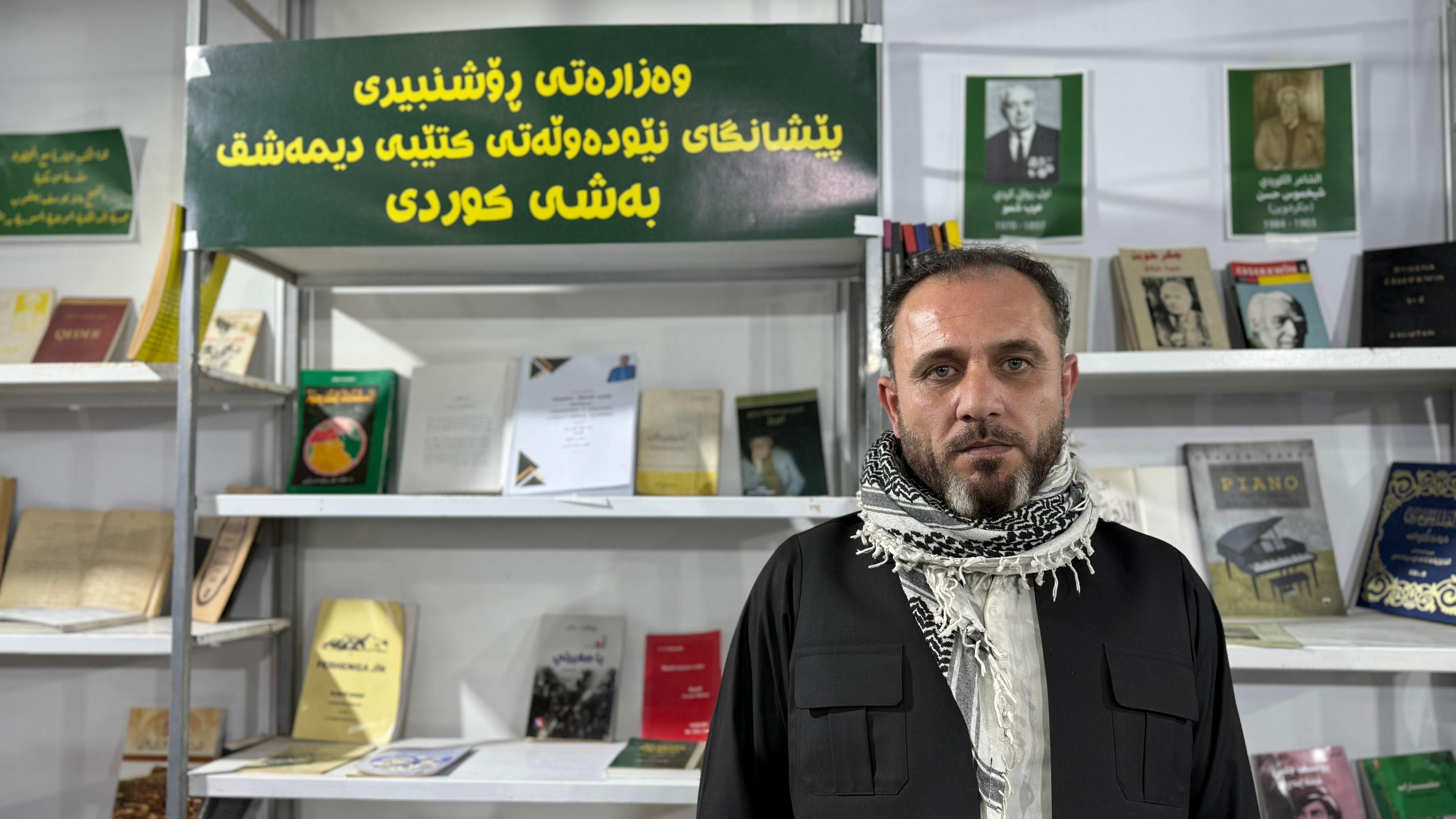 رجل ذو لحية وشارب يرتدي الوشاح الكردي التقليدي ويقف أمام أرفف وضع عليها كتب باللغة الكردية 