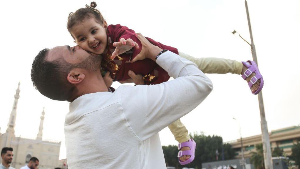 একজন লোক তার বাচ্চা মেয়েকে কোলে ধরে তার গালে চুমু খাচ্ছে।