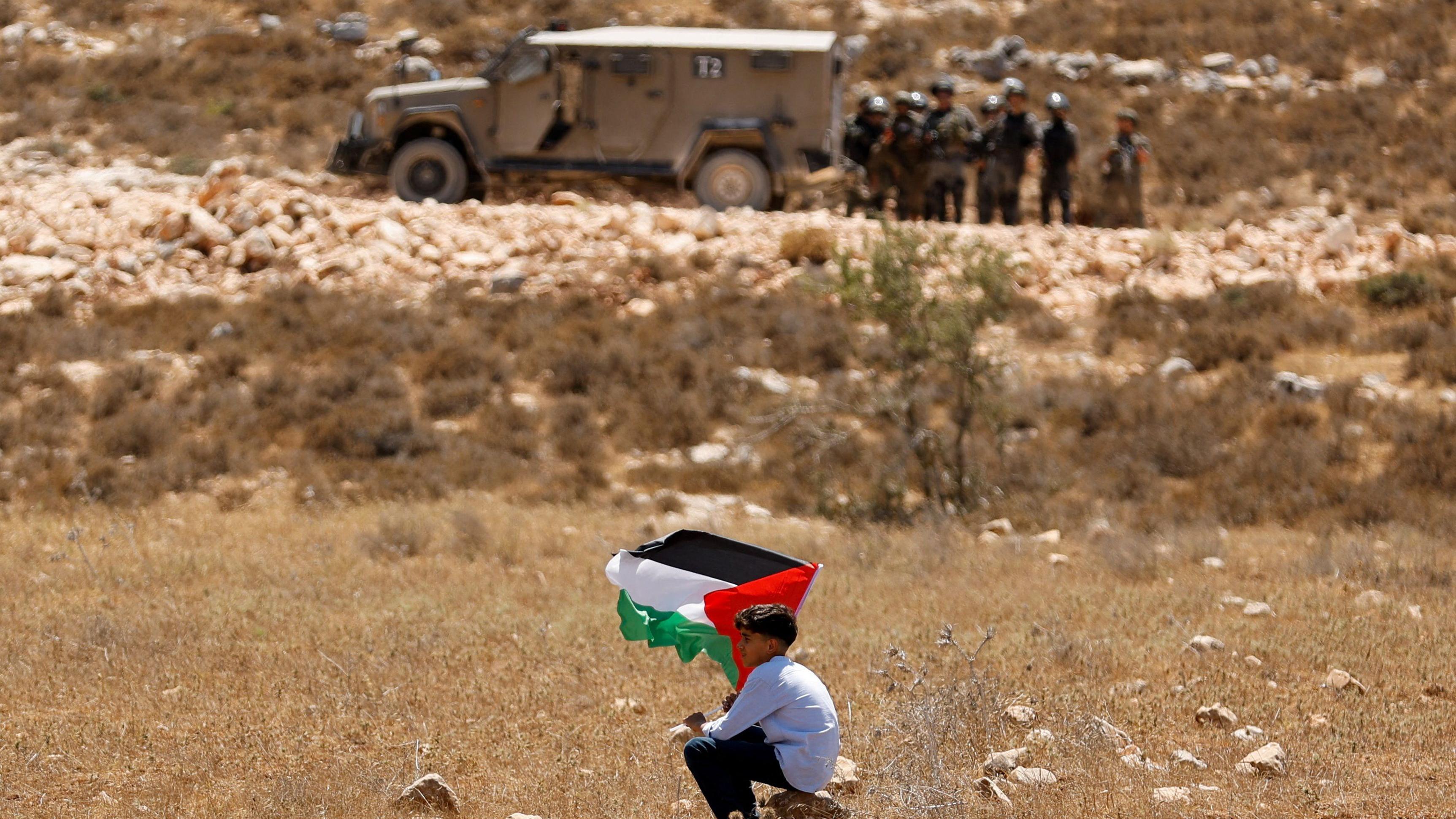 فتى يحمل العلم الفلسطيني خلال مظاهرة ضد ما يقول فلسطينيون إنه استيلاء إسرائيل على أراضيهم، بينما تقف قوات أمن إسرائيلية قرب جنين في الضفة الغربية