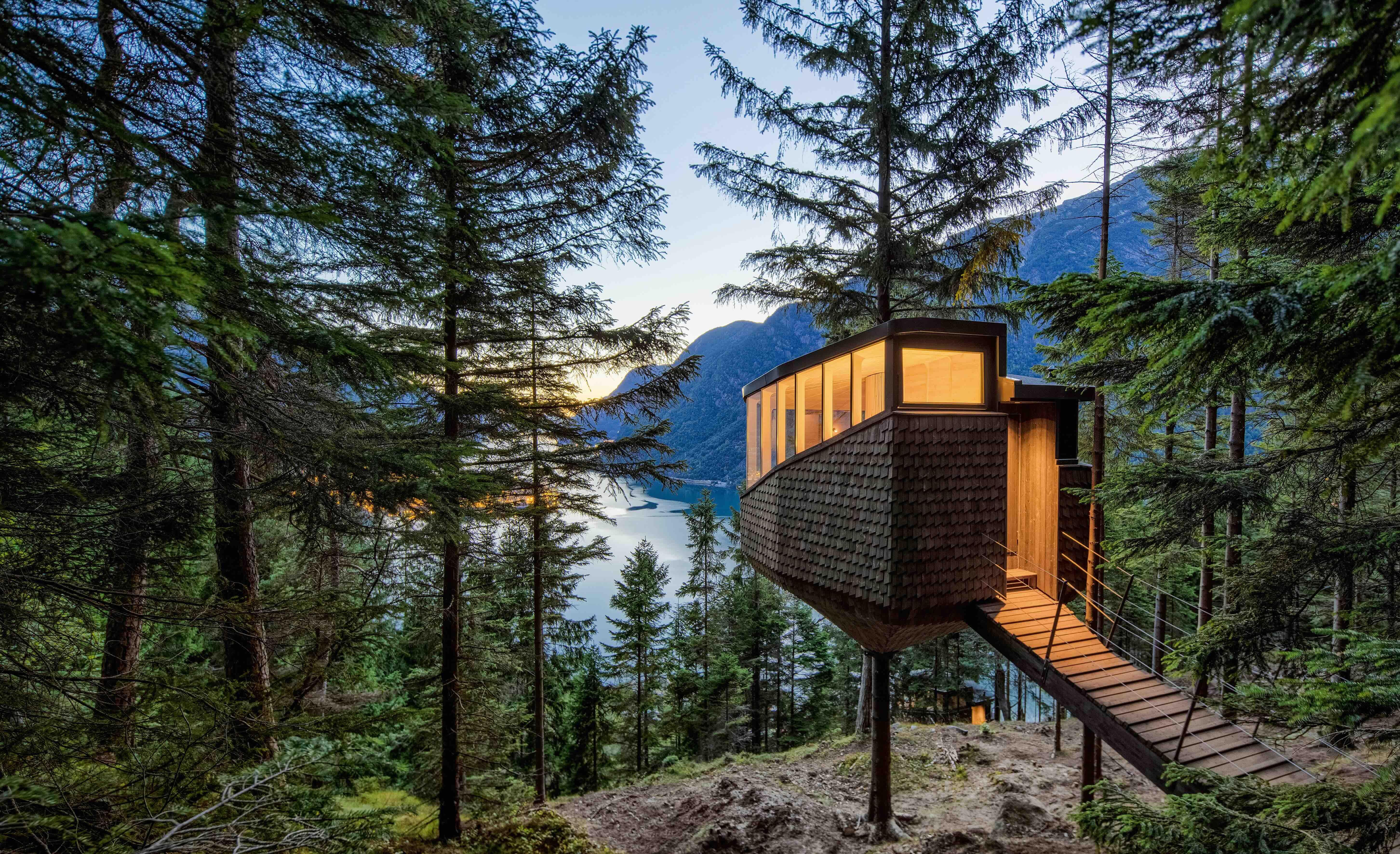 Una cabaña rectangular se erige sobre una loma que da a un lago y dos montañas que surgen a lado y lado.