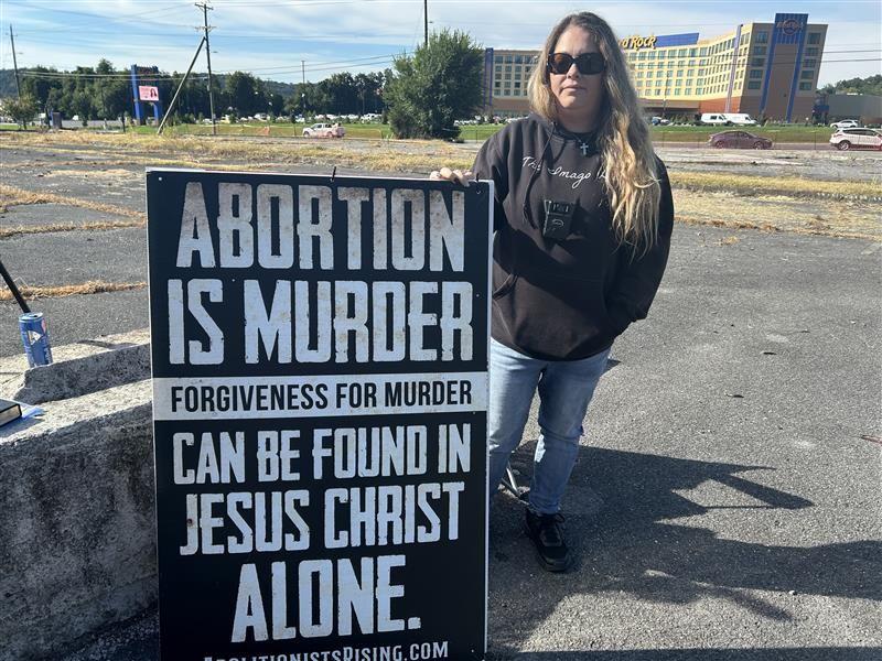 Uma mulher est&aacute; em p&eacute; em um estacionamento segurando um cartaz que diz: &ldquo;Aborto &eacute; assassinato. O perd&atilde;o pelo assassinato pode ser encontrado somente em Jesus Cristo&rdquo;. 