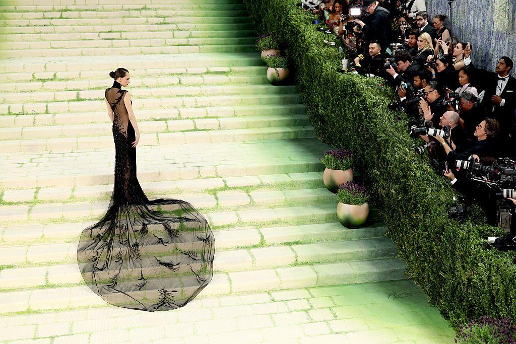 Una persona posa sola en una amplia escalinata verde durante un evento de gala, con un vestido negro con larga cola transparente, mientras fotógrafos capturan imágenes desde un lateral.
