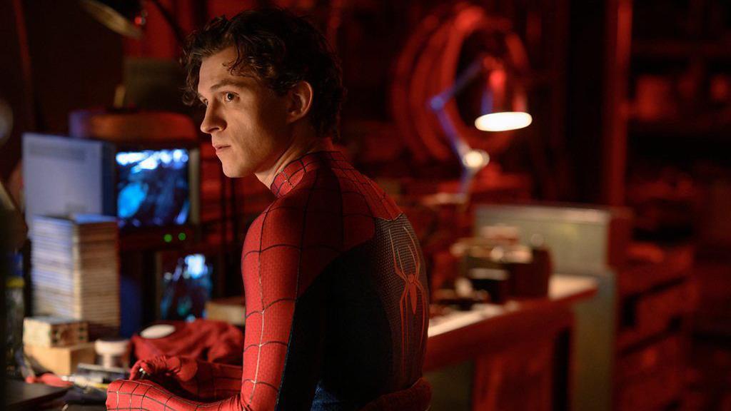 Tom Holland sem m&aacute;scara, com a roupa do Homem-Aranha, de costas, olhando para o lado em frente a uma mesa