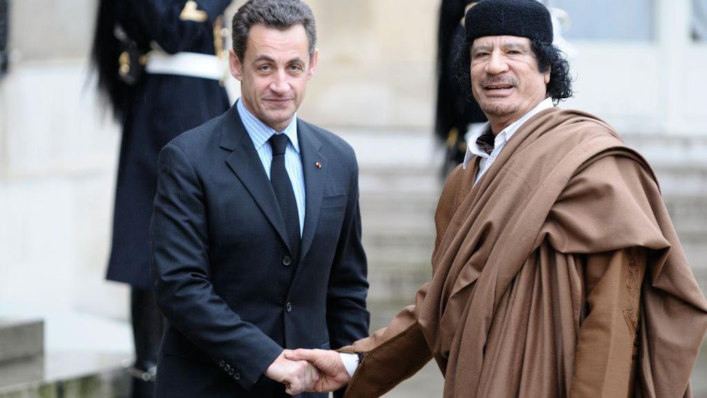 Nicolas Sarkozy, camisa de rayas celestes y blancas; corbata y chaqueta de traje oscuros, mira ala cámara mientras le da la mano derecha a Muamar Gadafi, quien viste una túnica marrón y un sombrero tradicional libio de color negro y redondeado.
