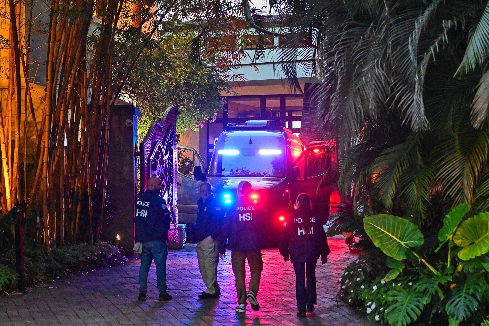 Policiais andam por uma construção à noite, com um carro de polícia piscando os faróis, rodeados pela vegetação e iluminados por diversas luzes