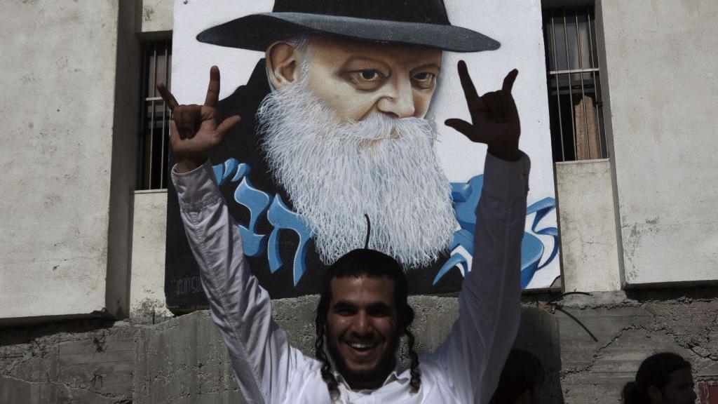 Homem erguendo os braços e sorrindo em frente a grafite com rosto de Menachem Mendel Schneerson
