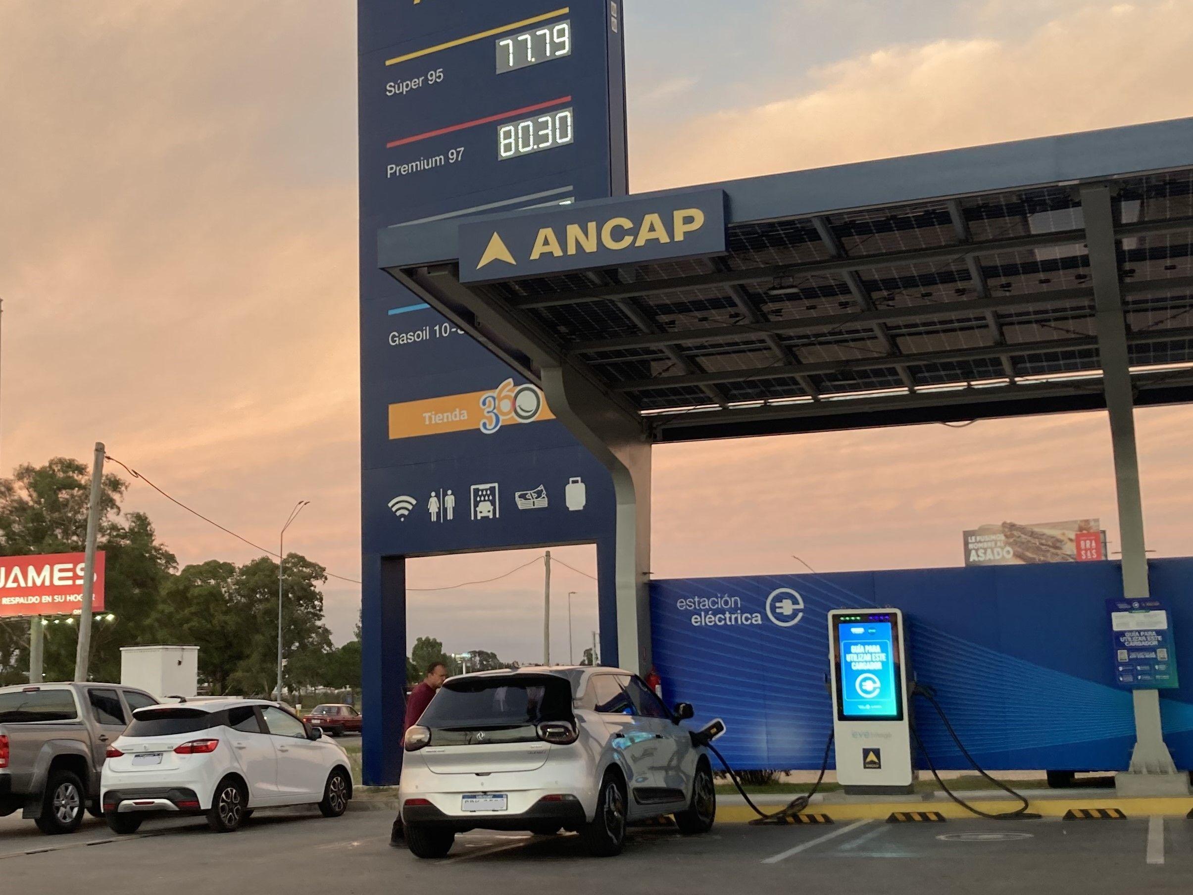 Un auto eléctrico carga su batería en la estación de una estación de servicio con precios de gasolina en un cartel.