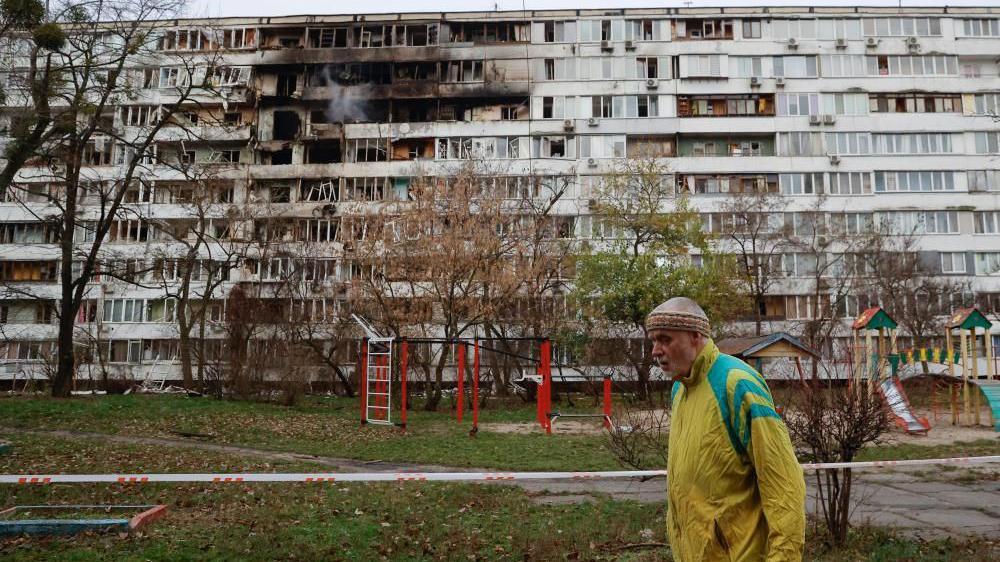 一名身穿黃色外套的男子走過一棟公寓大樓，建築表面明顯有空襲造成的損壞。
