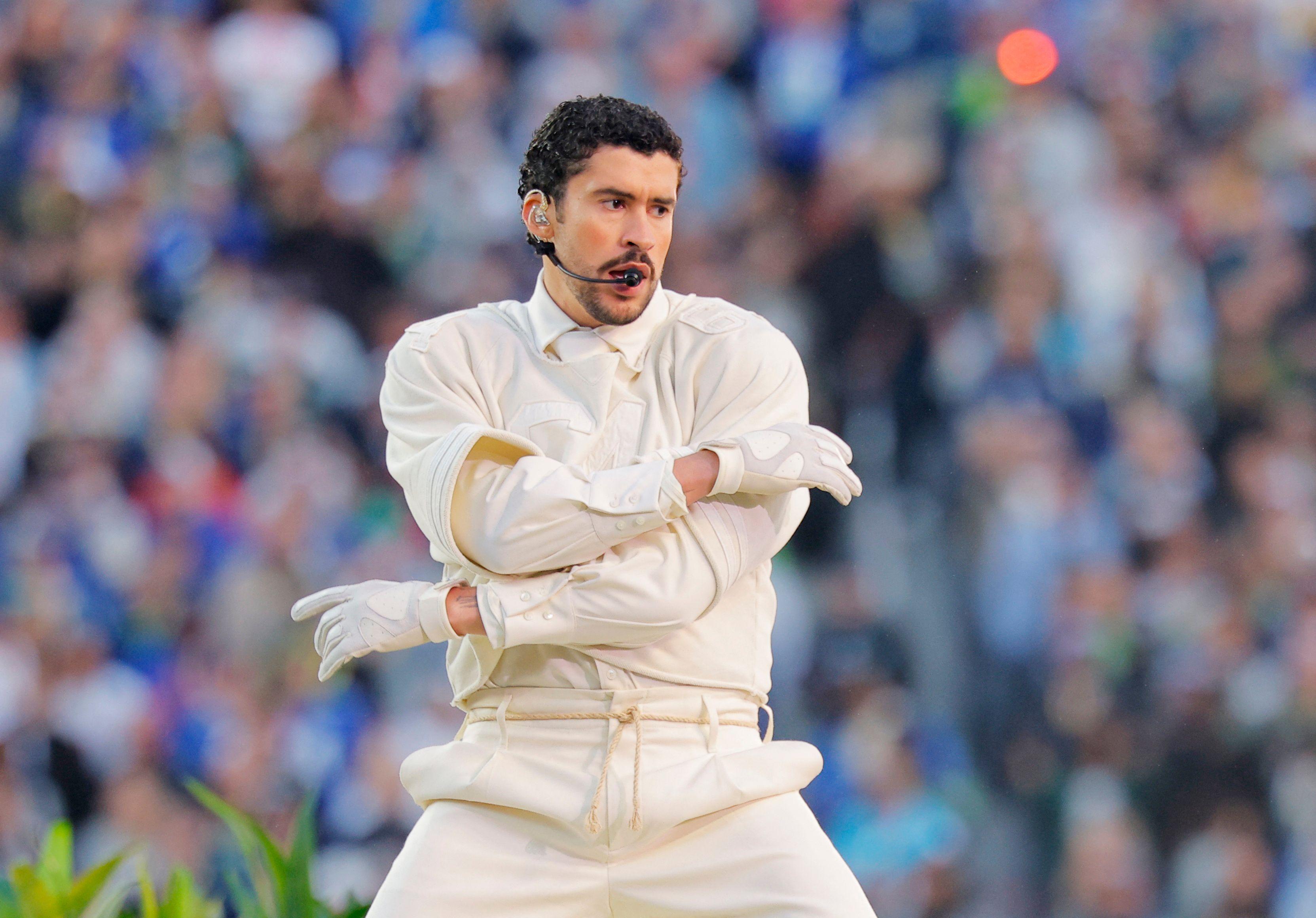 Homem se apresenta em um palco em um estádio, com um figurino bege e um microfone de cabeça. Ele está no centro da imagem, com os braços cruzados em movimento, expressão concentrada e boca entreaberta. Ao fundo, uma grande multidão aparece desfocada.