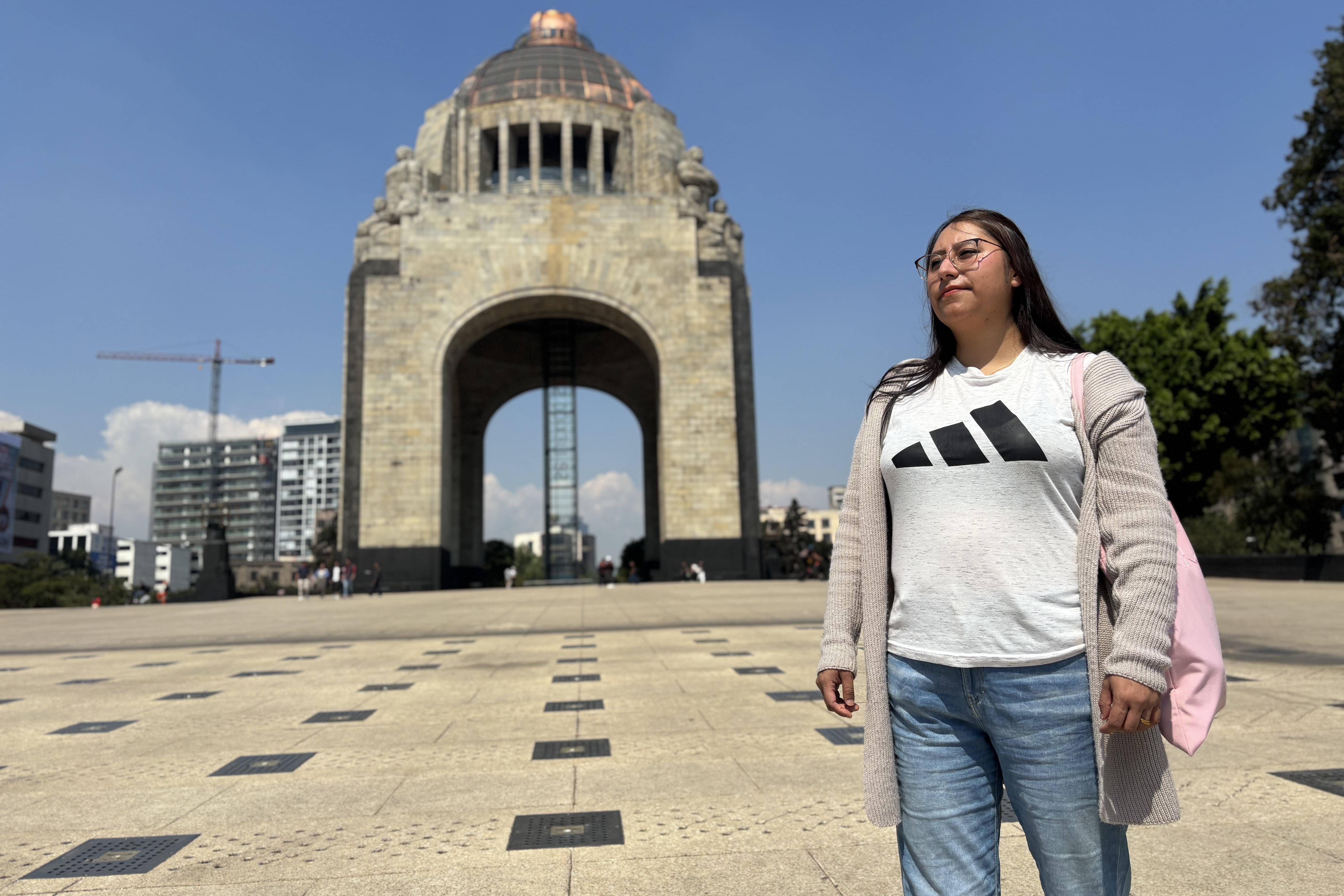 Mujer de pie en una plaza amplia frente al Monumento a la Revolución, con edificios urbanos al fondo y cielo despejado.