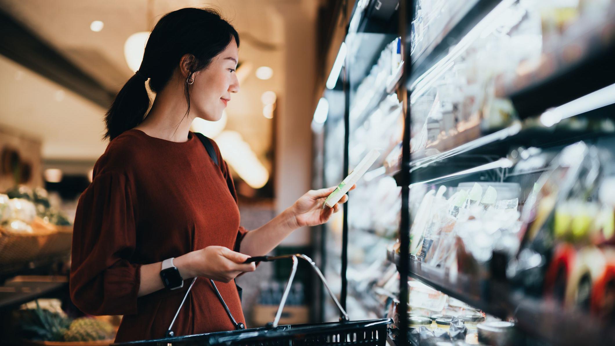 Una mujer observa un producto que lleva en la sección refrigerada de un supermercado. Lleva un vestido marrón y sostiene una cesta en una mano.