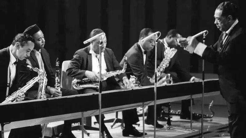 Djuk Elington u odelu diriguje orkestrom u prvim redovima nekoliko muzičara sa saksofonima