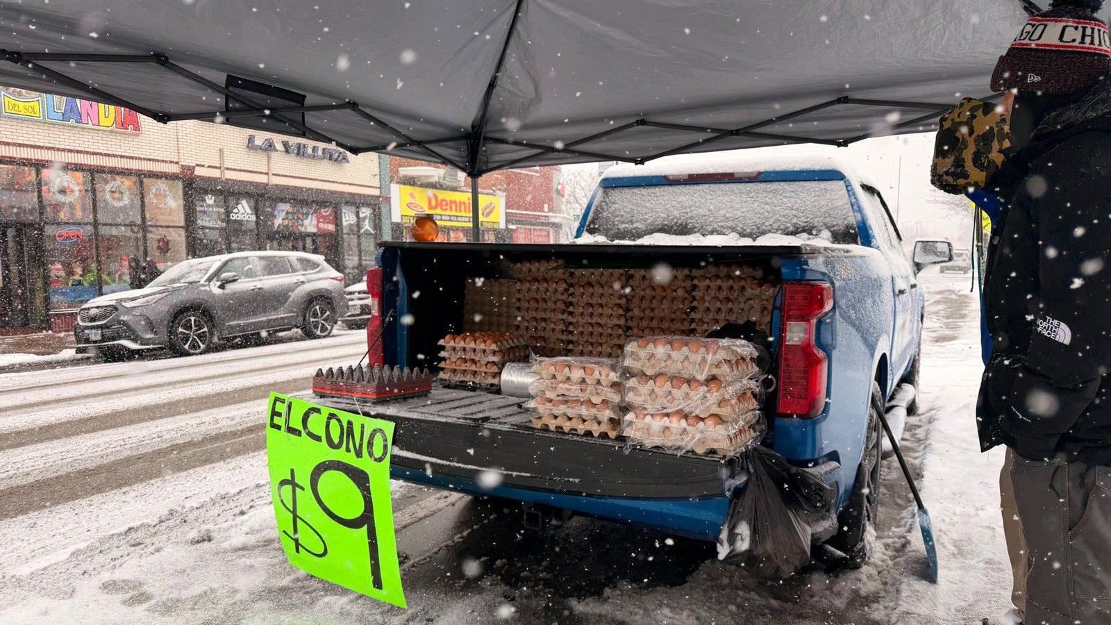 Un vendedor ambulante vende huevos desde una camioneta azul estacionada en una calle donde ha caído una nevada. Un letrero verde anuncia: El cono, $9