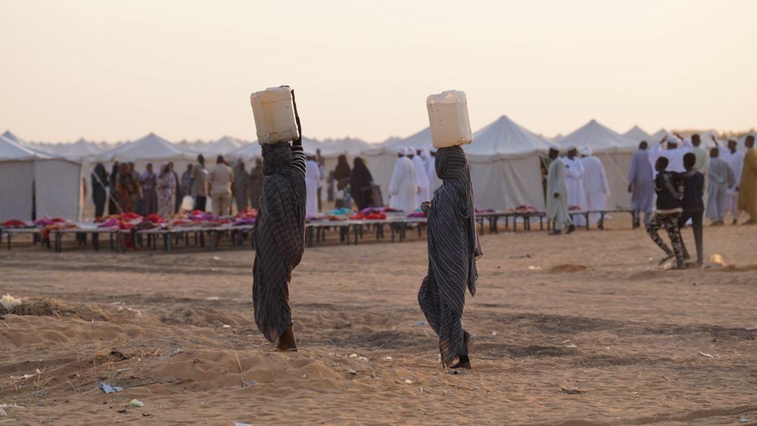 سيدتان تسيران أمام الخيم وتحملان أوعية بلاستيكية على رأسيهما 