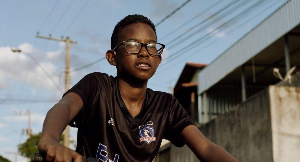 Um adolescente negro, o ator C&iacute;cero Lucas, usa &oacute;culos e pedala uma bicicleta em uma rua de bairro residencial, sob a luz dourada do fim da tarde. Ele veste uma camisa preta de futebol e olha para frente com express&atilde;o concentrada, enquanto casas, postes e fios el&eacute;tricos aparecem ao fundo.