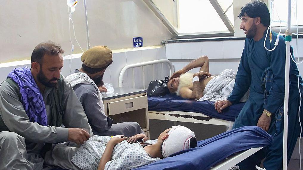 Un niño con una venda en la cabeza y un anciano, ambos tendidos en camas, reciben atención médica en un hospital, rodeados de tres adultos