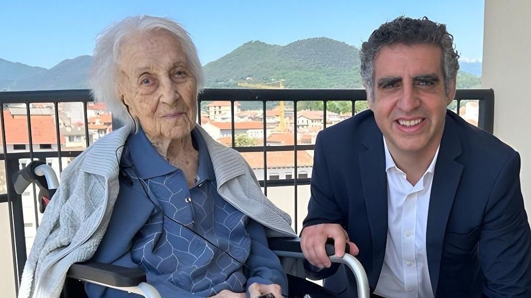 María Branyas sentada en una silla de ruedas, con un saco azul, en un balcón posa junto al doctor Manel Esteller