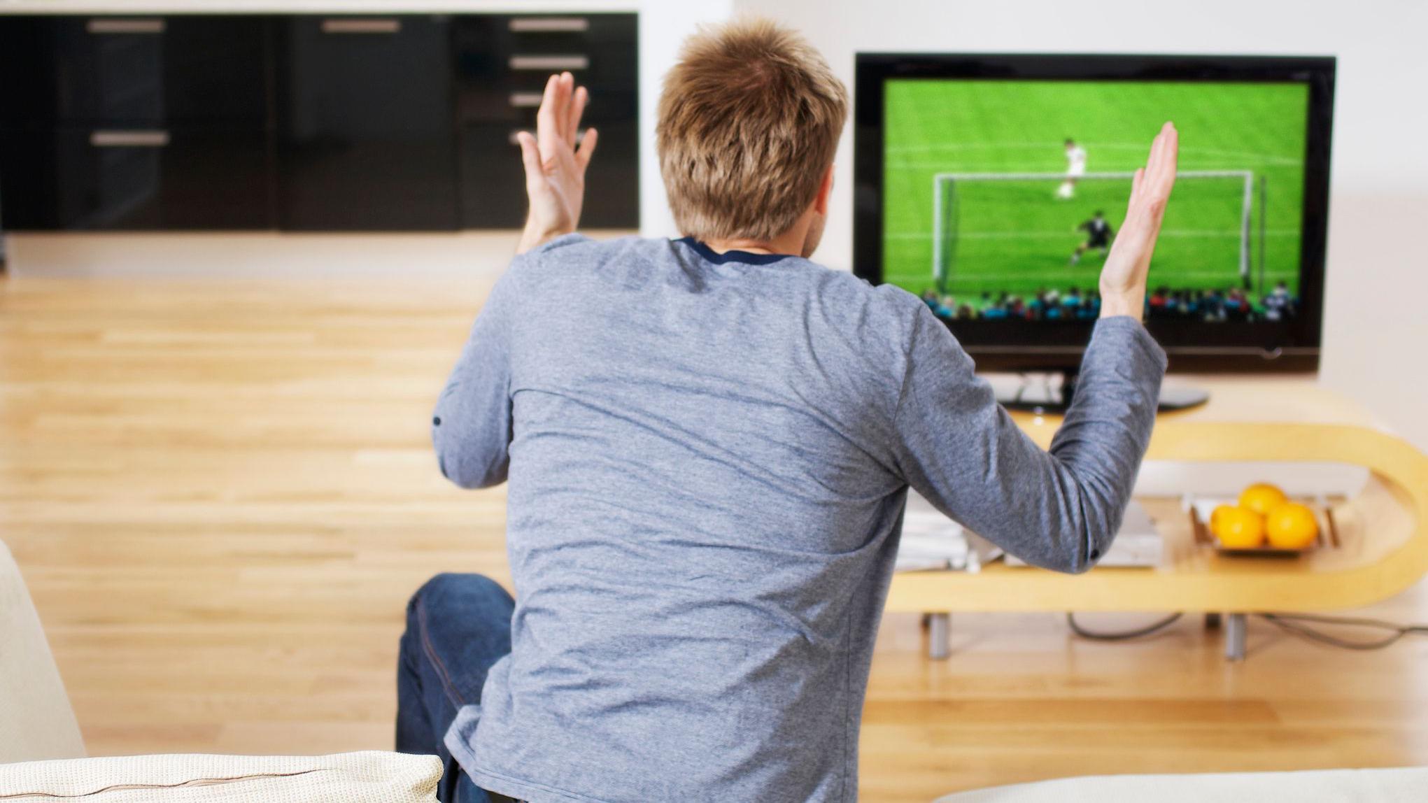 شاب من الخلف يشاهد مباراة كرة قدم على الشاشة ويرفع يديه متفاعلاً