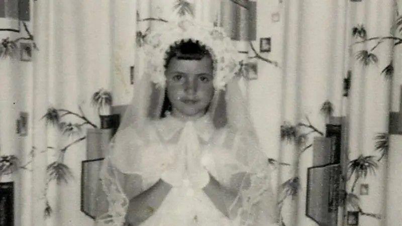 Foto preta e branca que mostra uma criança usando uma roupa branca, um véu e juntando as duas mãos em oração