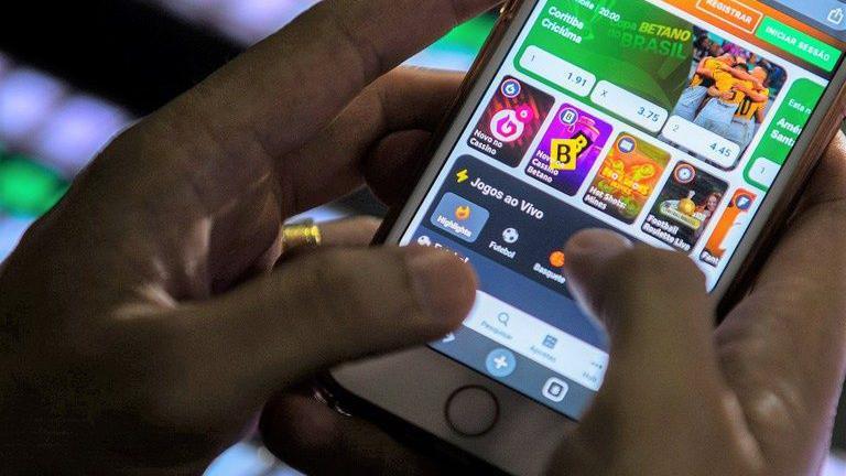 M&atilde;os segurando um celular que mostra na tela &iacute;cones de sites de apostas