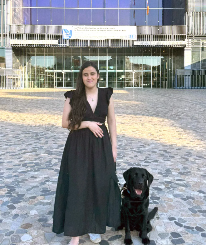 Una mujer con vestido negro, de pie junto a un perro lazarillo negro. 