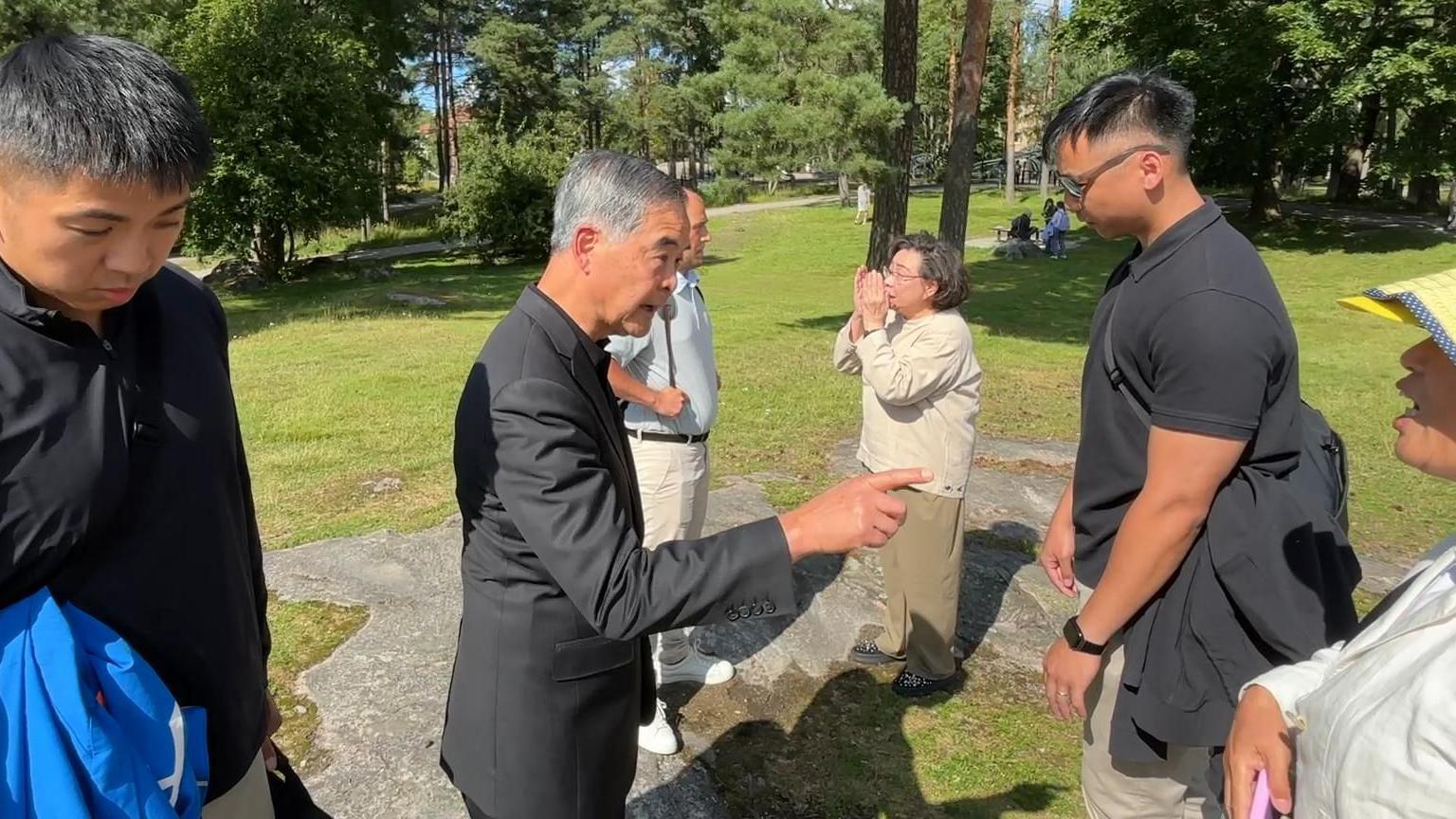 芬蘭赫爾辛基西貝柳斯公園內，身穿黑色西裝的梁振英（中）指罵一名頭戴淺黃色帽子的女性法輪功學員，身穿米白色上衣的夫人梁唐青儀（右三）與一名歐洲男子交談，兩名身穿黑色上衣的疑似保鑣站在梁振英左右兩側垂下頭來（法輪大法信息中心發放影片截屏13/8/2025）