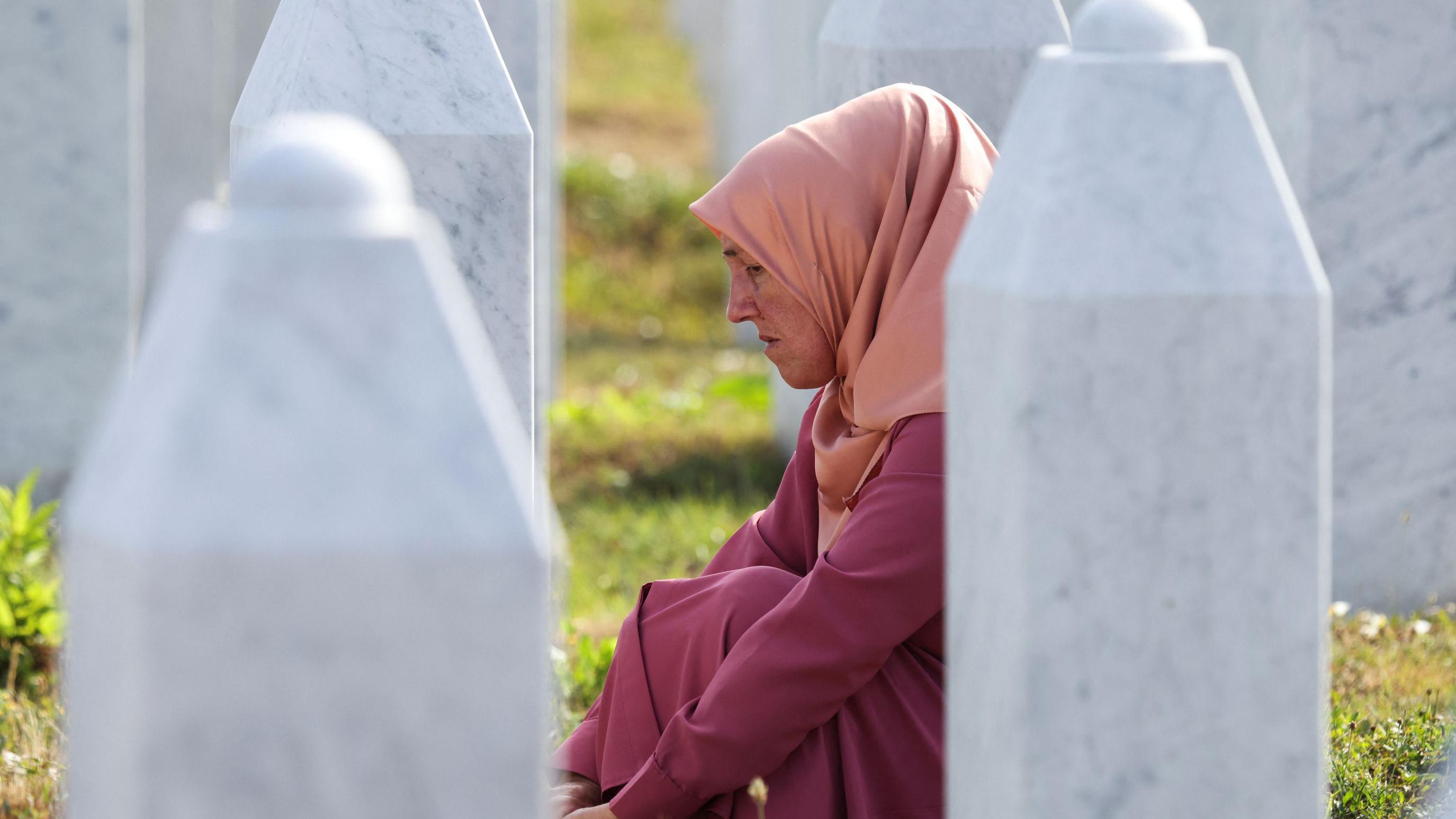 Srebrenica i genocid: Jul 1995. koji se ne zaboravlja 1 Žena prekrivene glave pognute glave sedi na travi između nadgrobnih spomenika u Potočarima